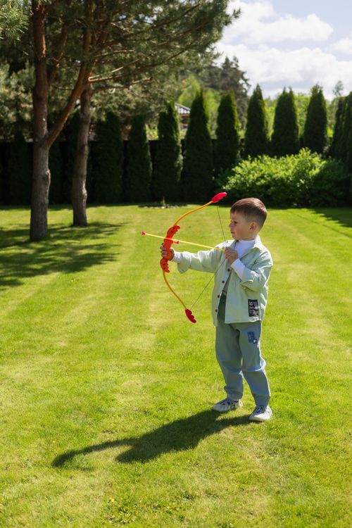 Free A child enjoys playing outdoors with a toy bow and arrow, surrounded by green grass and trees. Stock Photo