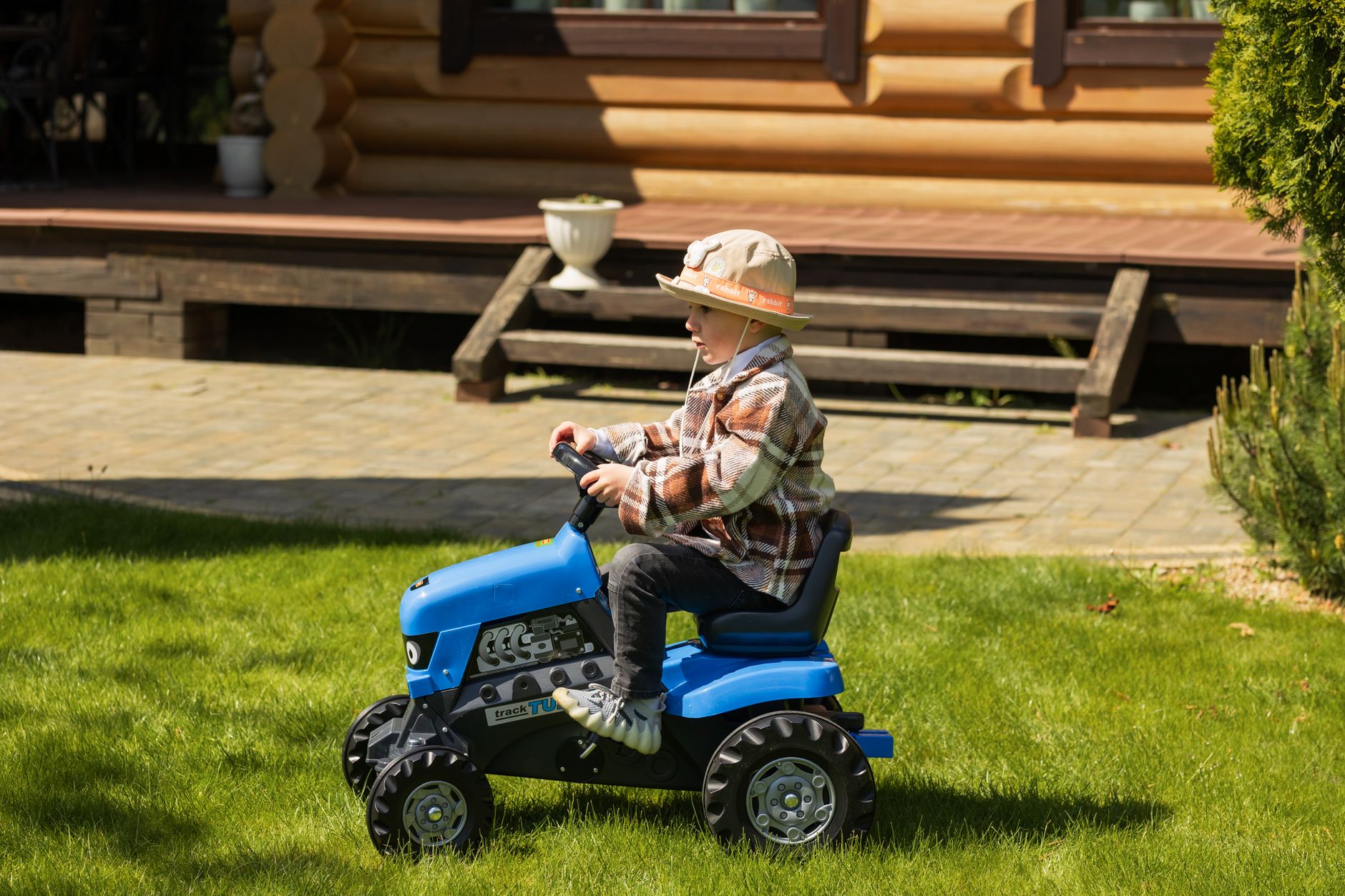 Young boy rides a blue toy tractor in a sunny garden near a wooden cabin.