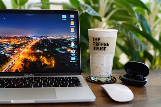Stylish workspace featuring a laptop, coffee glass, and earbuds against a leafy background.