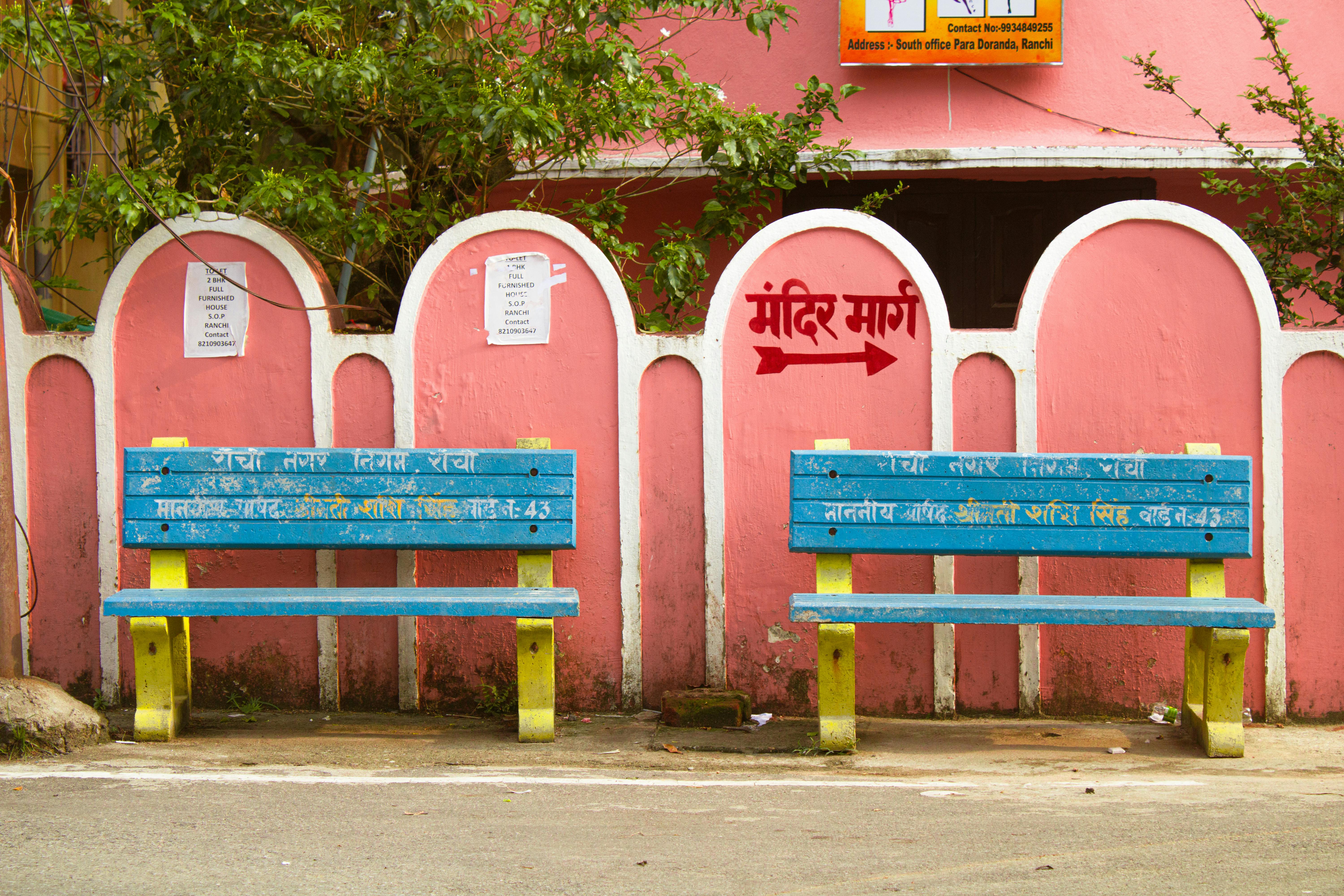 Gratis Dua bangku biru cerah berlatar dinding merah muda dengan tulisan-tulisan berbahasa Hindi di suasana perkotaan. Foto Stok