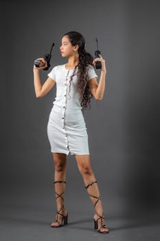 Young woman in a white dress holding walkie-talkies with a confident pose. Studio portrait on gray background.