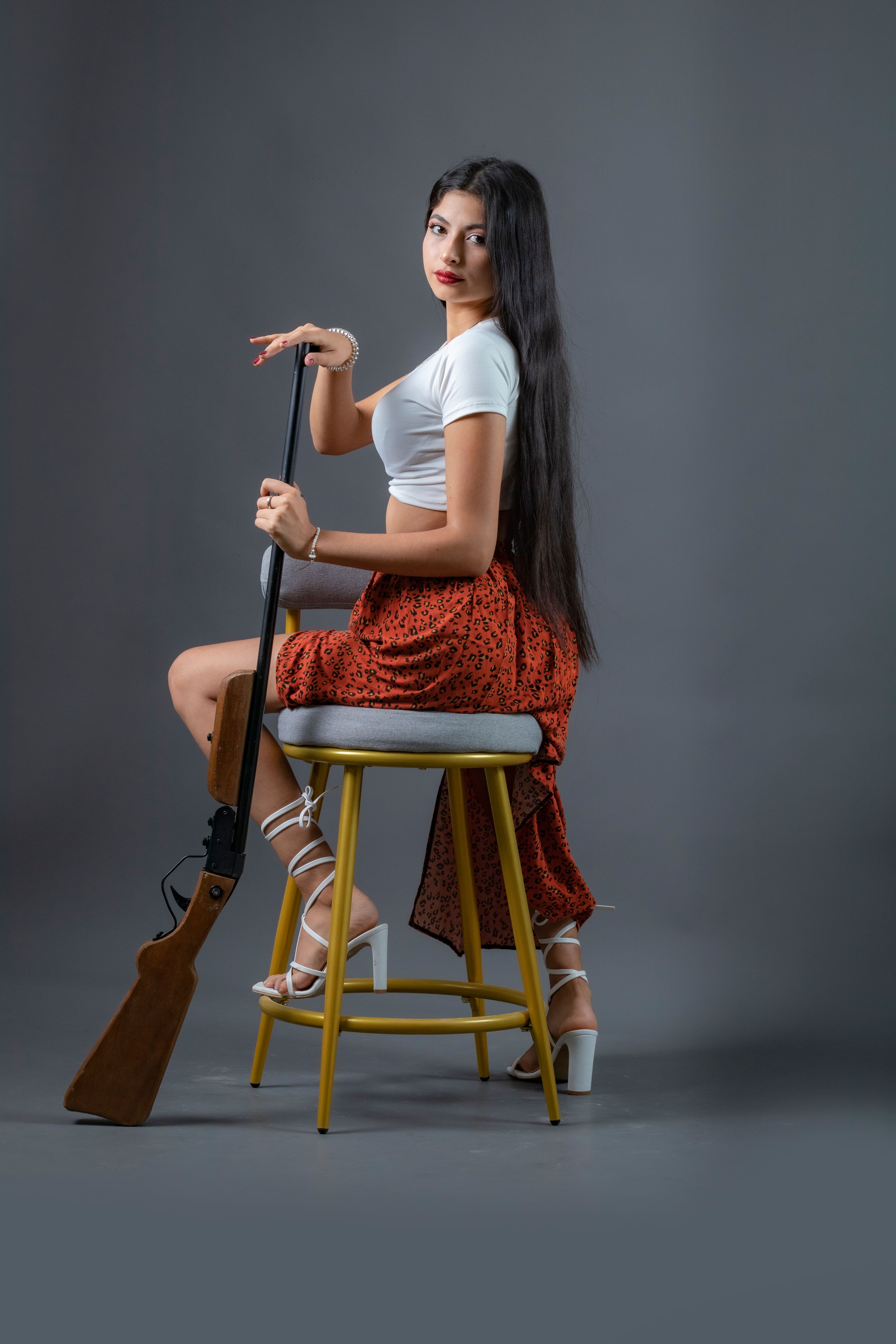 Woman sitting on stool holding rifle against a neutral background.