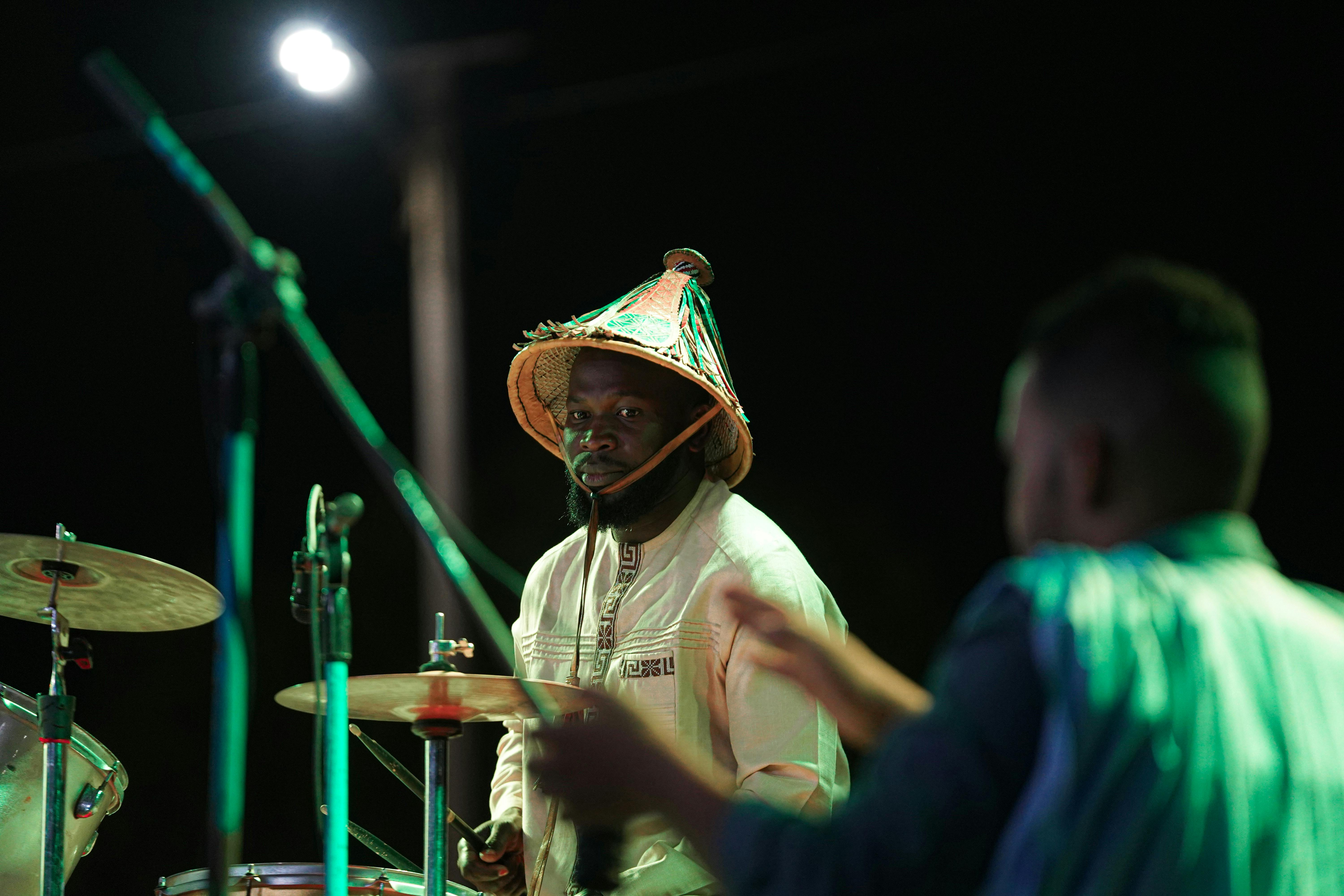 A drummer performs at an outdoor night concert wearing a traditional hat.