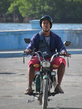 A man on motorcycle by a waterfront, wearing a helmet and casual clothing.