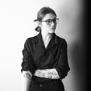 A black and white portrait featuring a woman with glasses, tattoos, and a pensive expression.