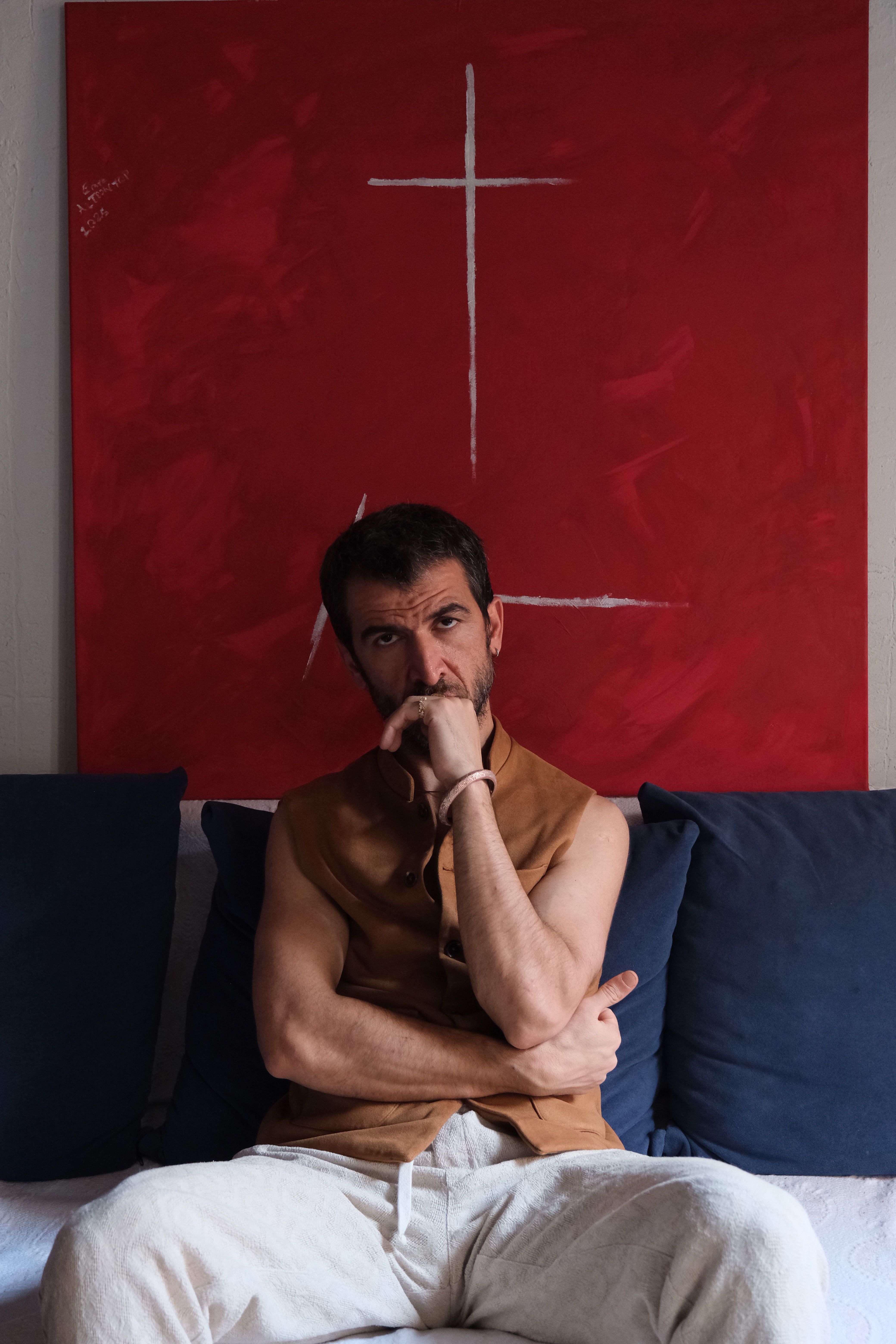 A man thoughtfully sits against a bold red abstract painting indoors.