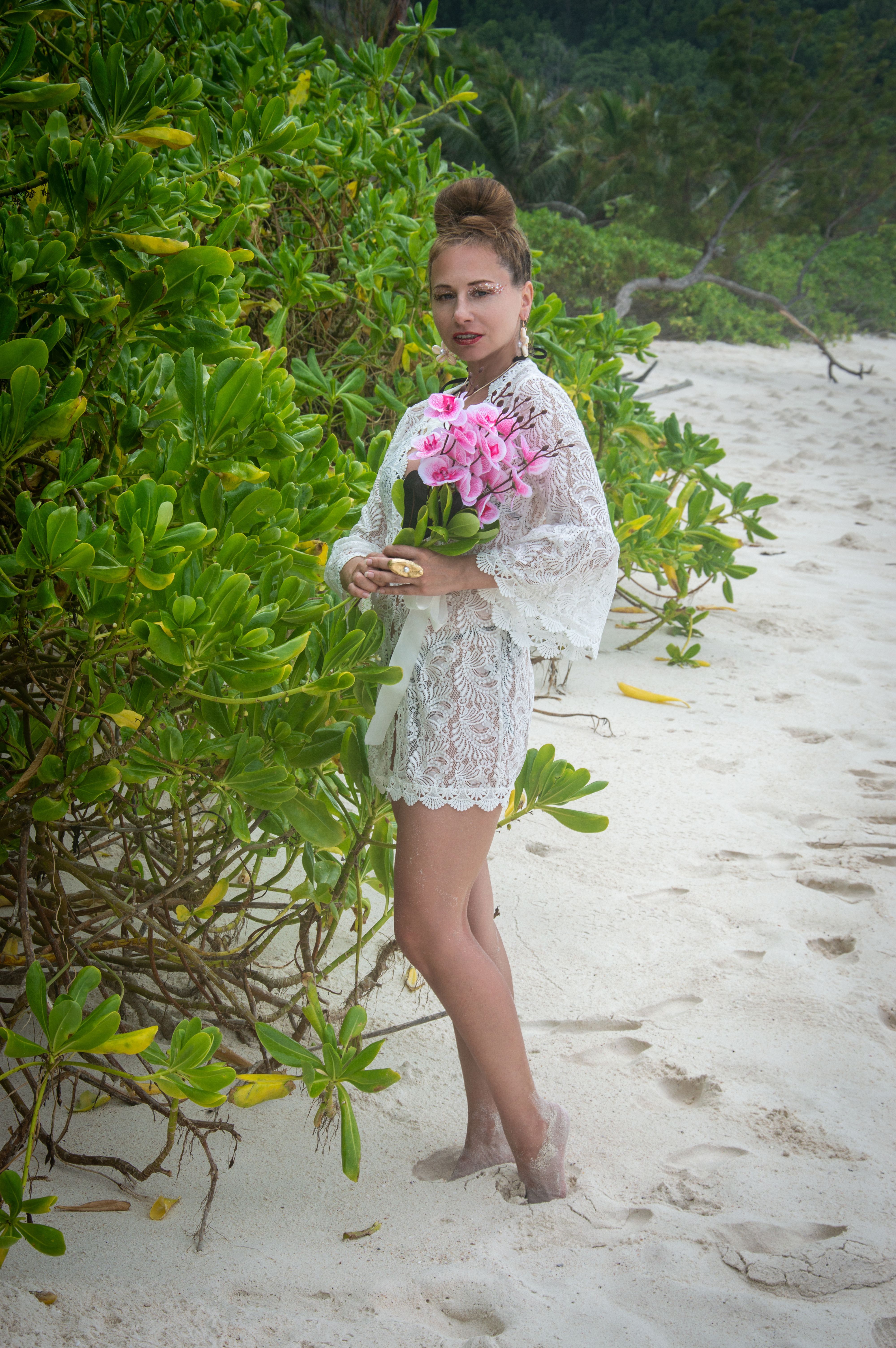 A woman in lace dress holding pink lilies stands on a tropical beach surrounded by lush greenery.