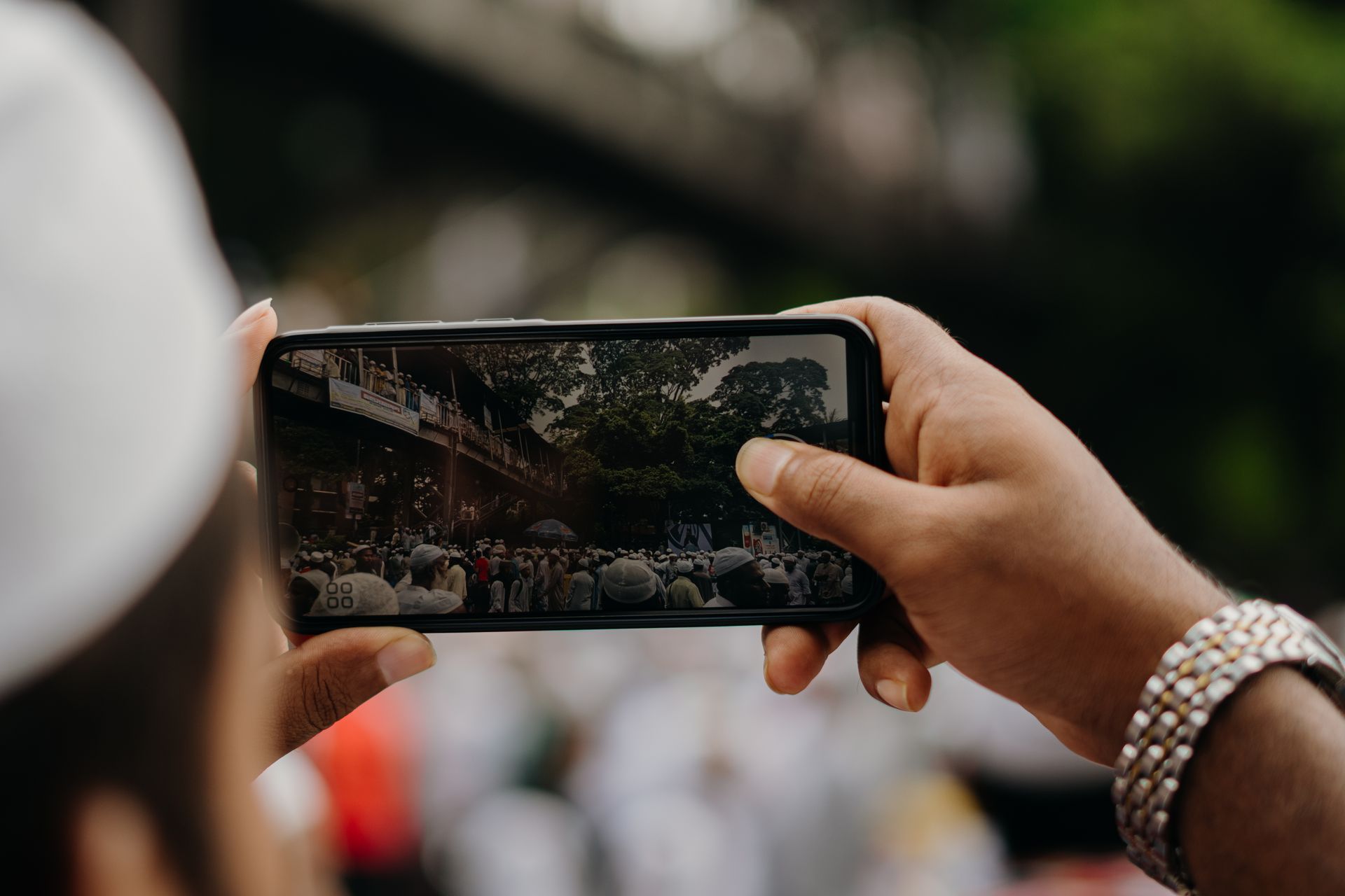 Close-up of hands holding a smartphone, capturing a lively outdoor event with a crowd of people.