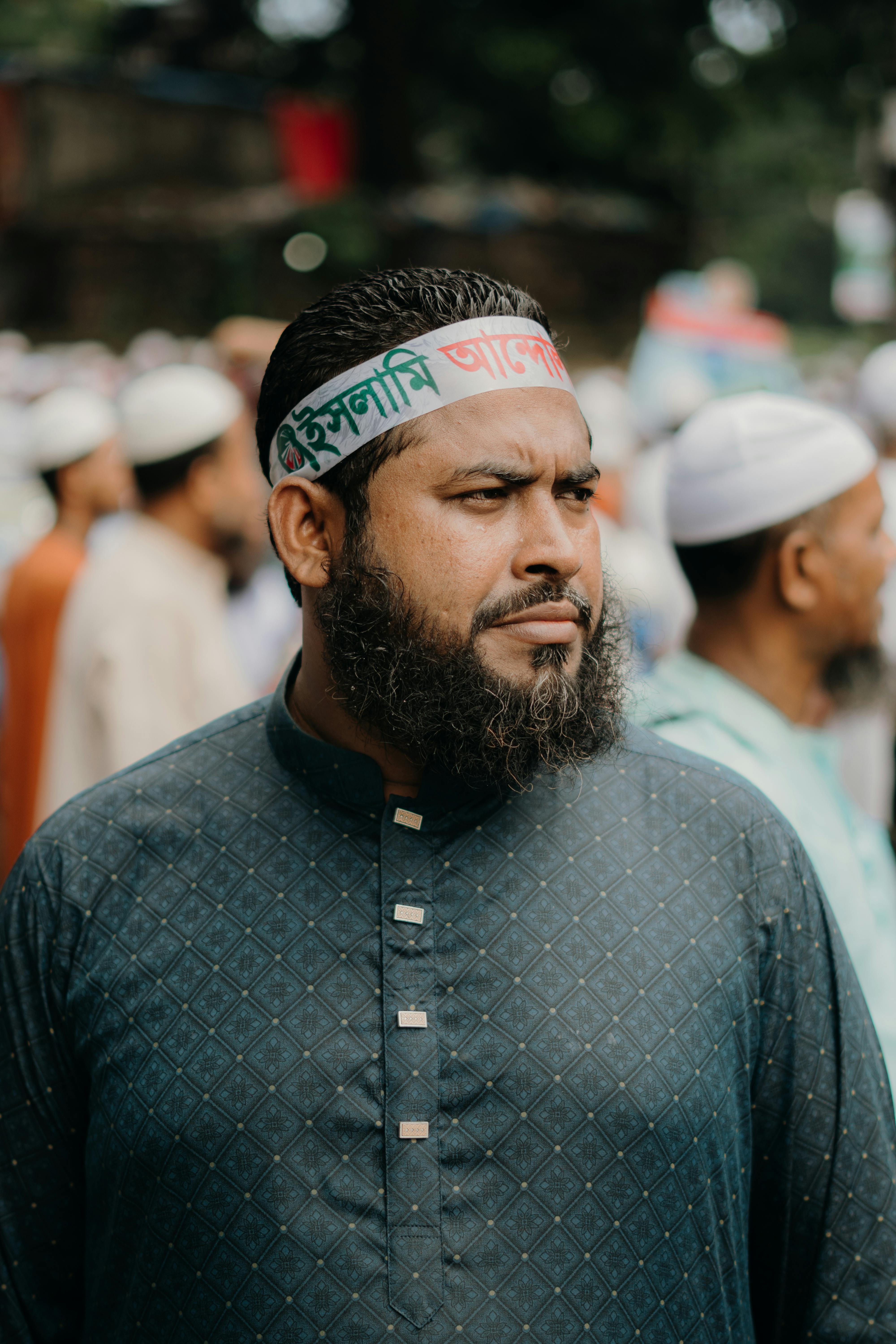 Bearded man wearing traditional attire with a headband at an outdoor gathering, focused expression.