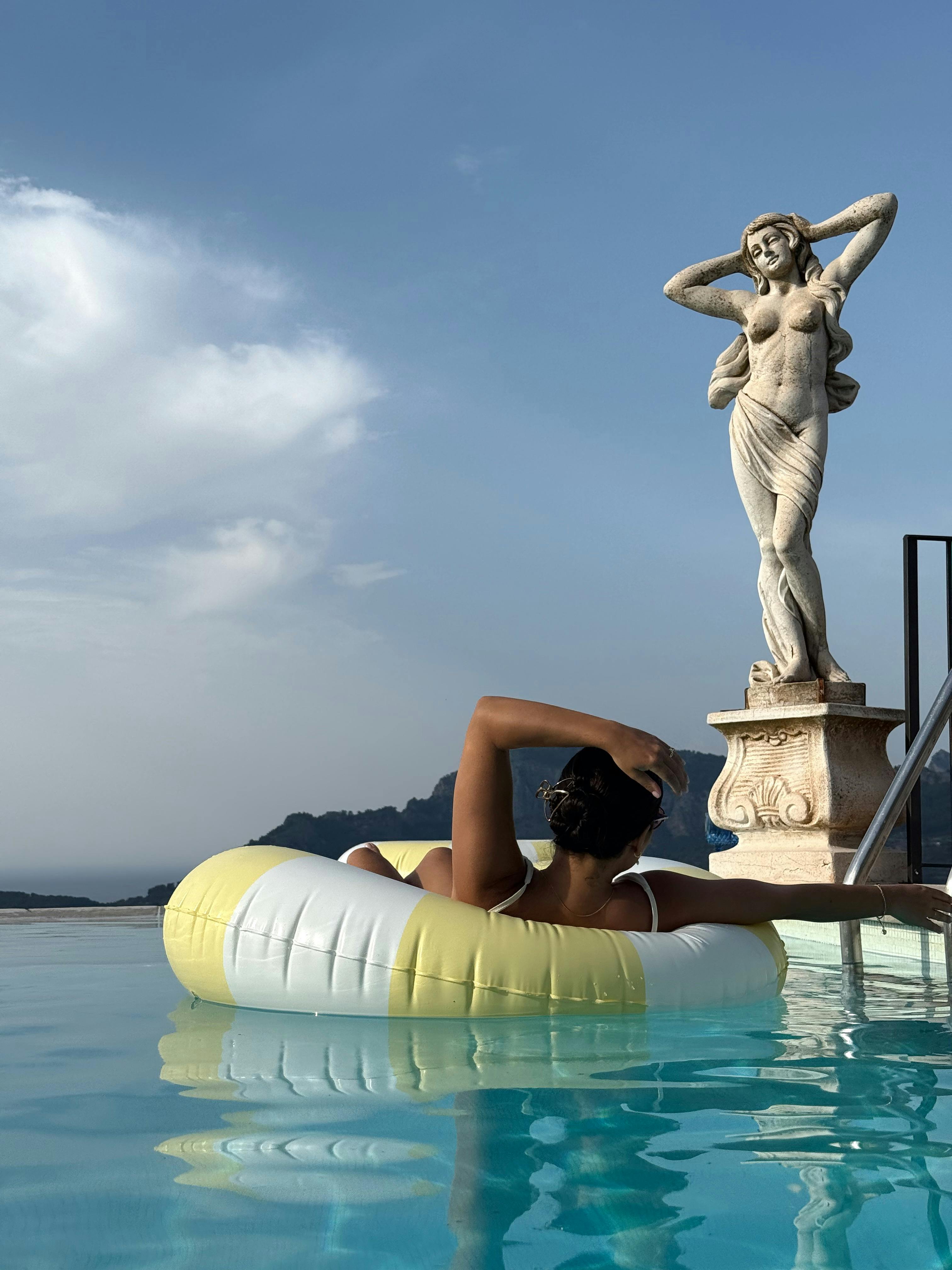 Free Woman enjoying a leisure float in a pool beside a classical statue under a clear sky. Stock Photo