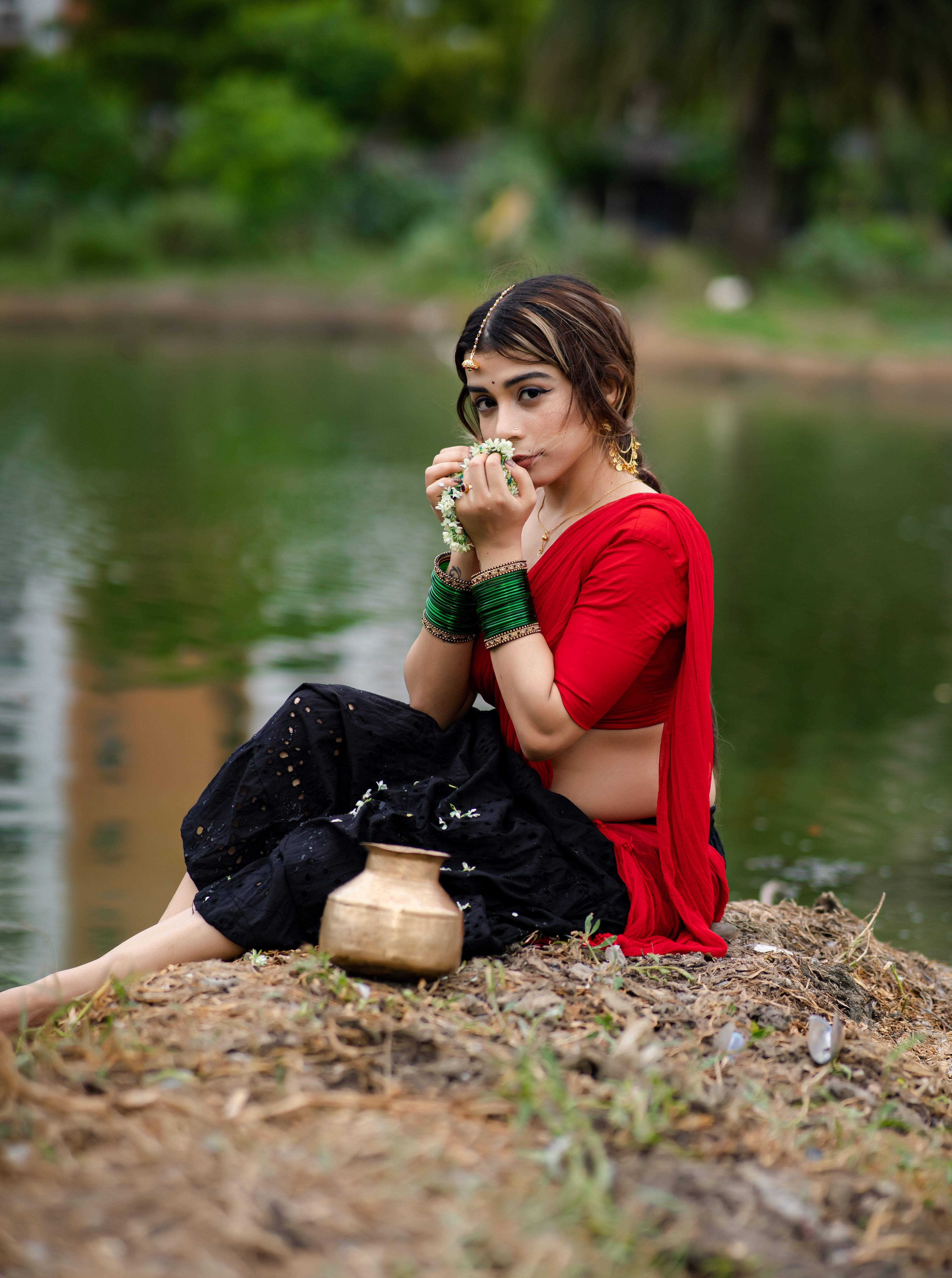 Woman in traditional attire by a river, holding flowers in a serene outdoor setting.