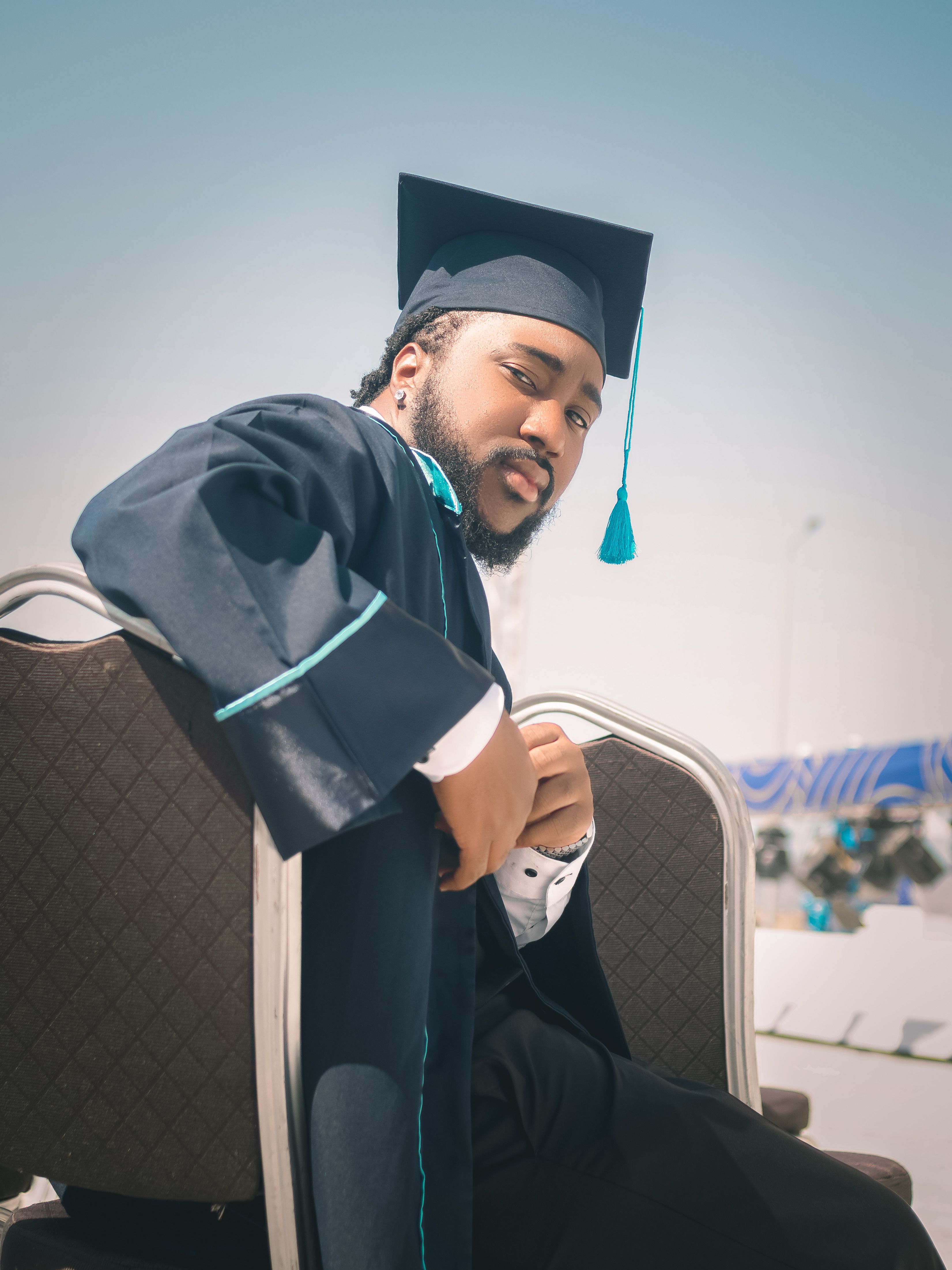 A proud graduate in cap and gown sits outdoors at a sunny graduation ceremony.