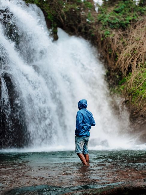 Man in blue jacket stands by a waterfall in Kerala, India, embracing nature's beauty.