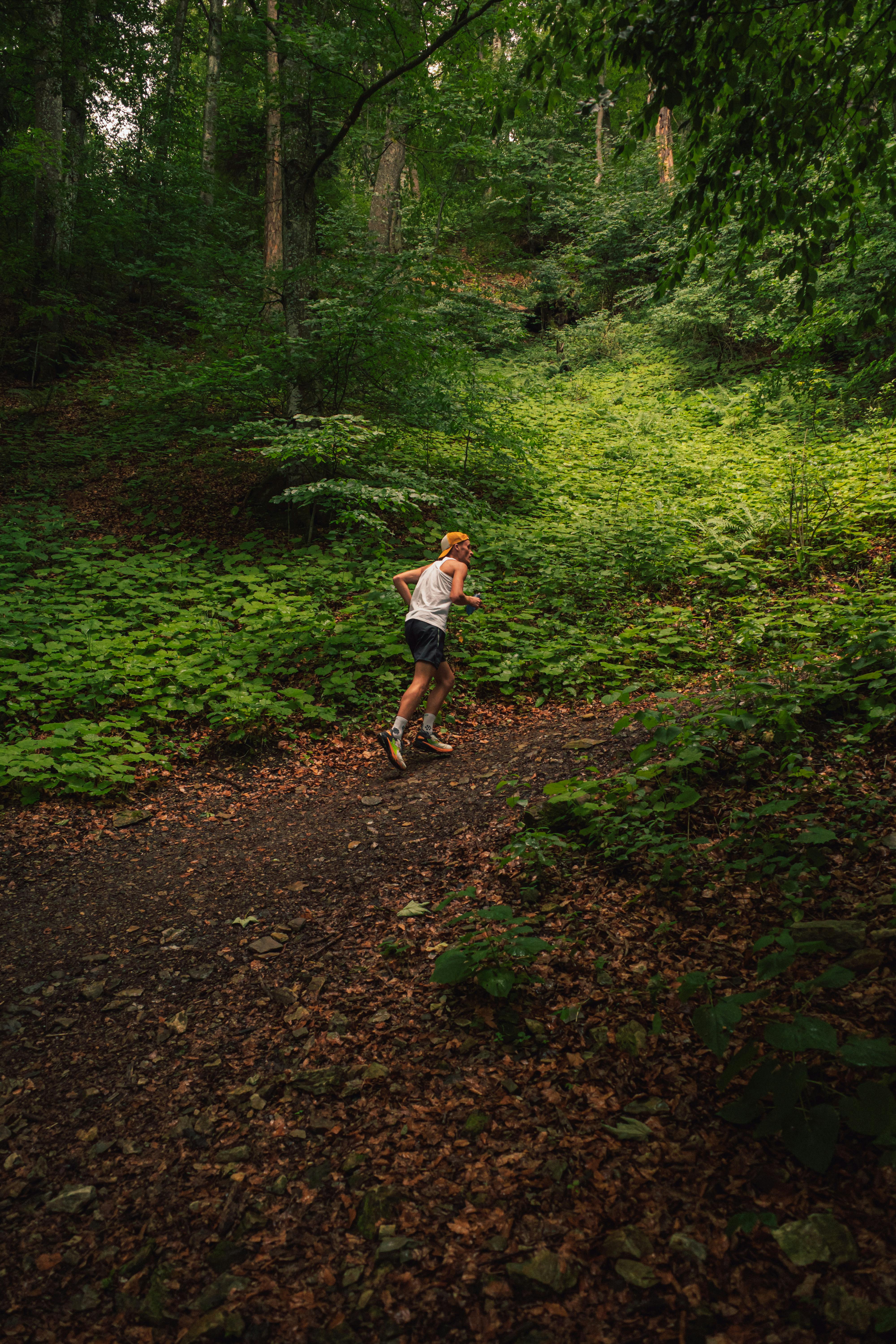 An athletic blonde teenager running on a forest trail surrounded by lush greenery and trees.