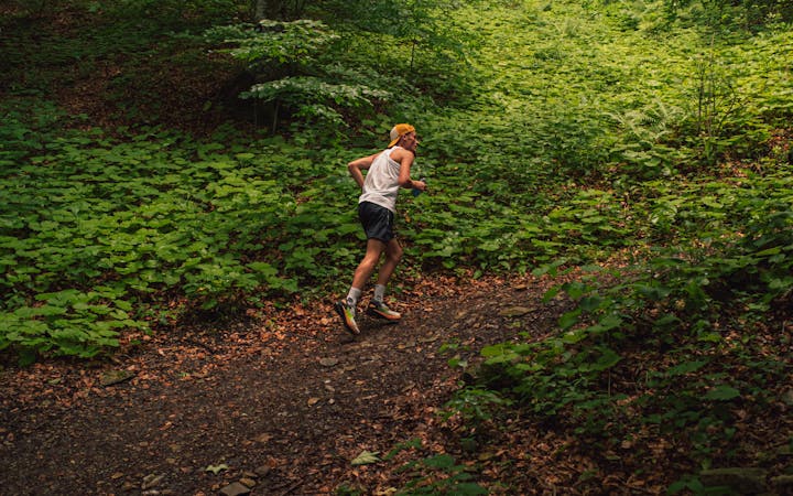 An athletic blonde teenager running on a forest trail surrounded by lush greenery and trees.