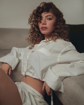 Woman with curly hair in a white casual outfit lounging indoors on a sofa.