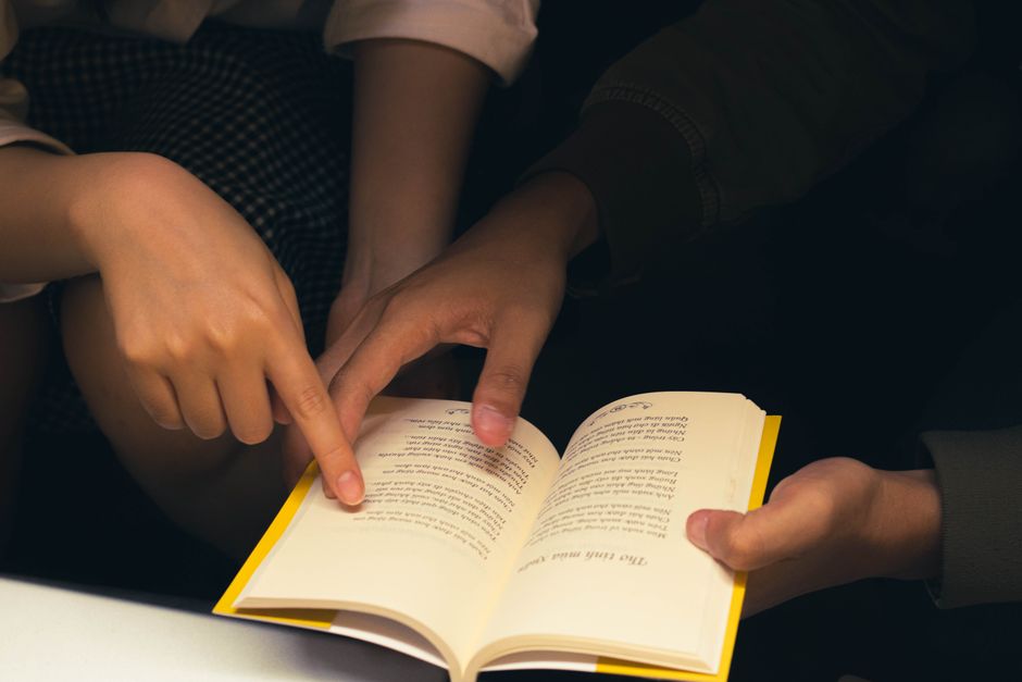 Close-up of two people sharing a book, pointing at text, creating a cozy reading atmosphere.