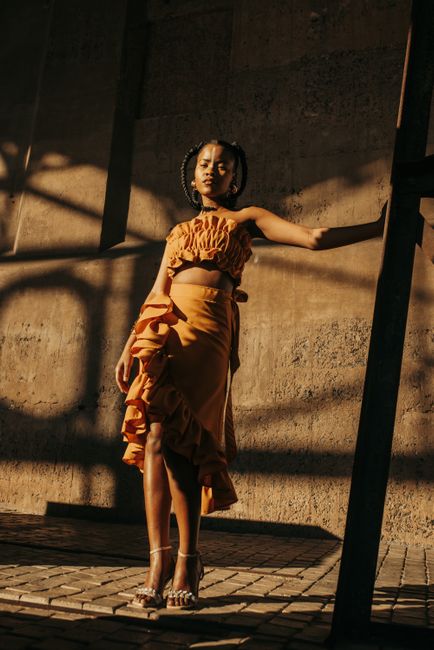 Stylish woman in a ruffled dress poses dramatically in warm light against a textured urban backdrop.