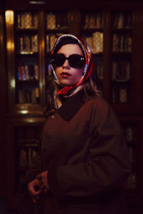 Free Elegant woman in sunglasses and vintage scarf stands in a library, exuding classic style and sophistication. Stock Photo