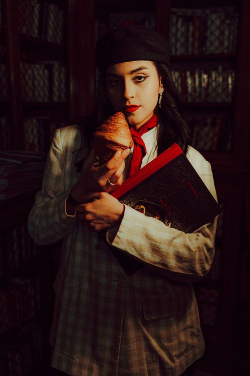 Free Stylish woman in library with a croissant and decorative book, exuding Parisian chic. Stock Photo