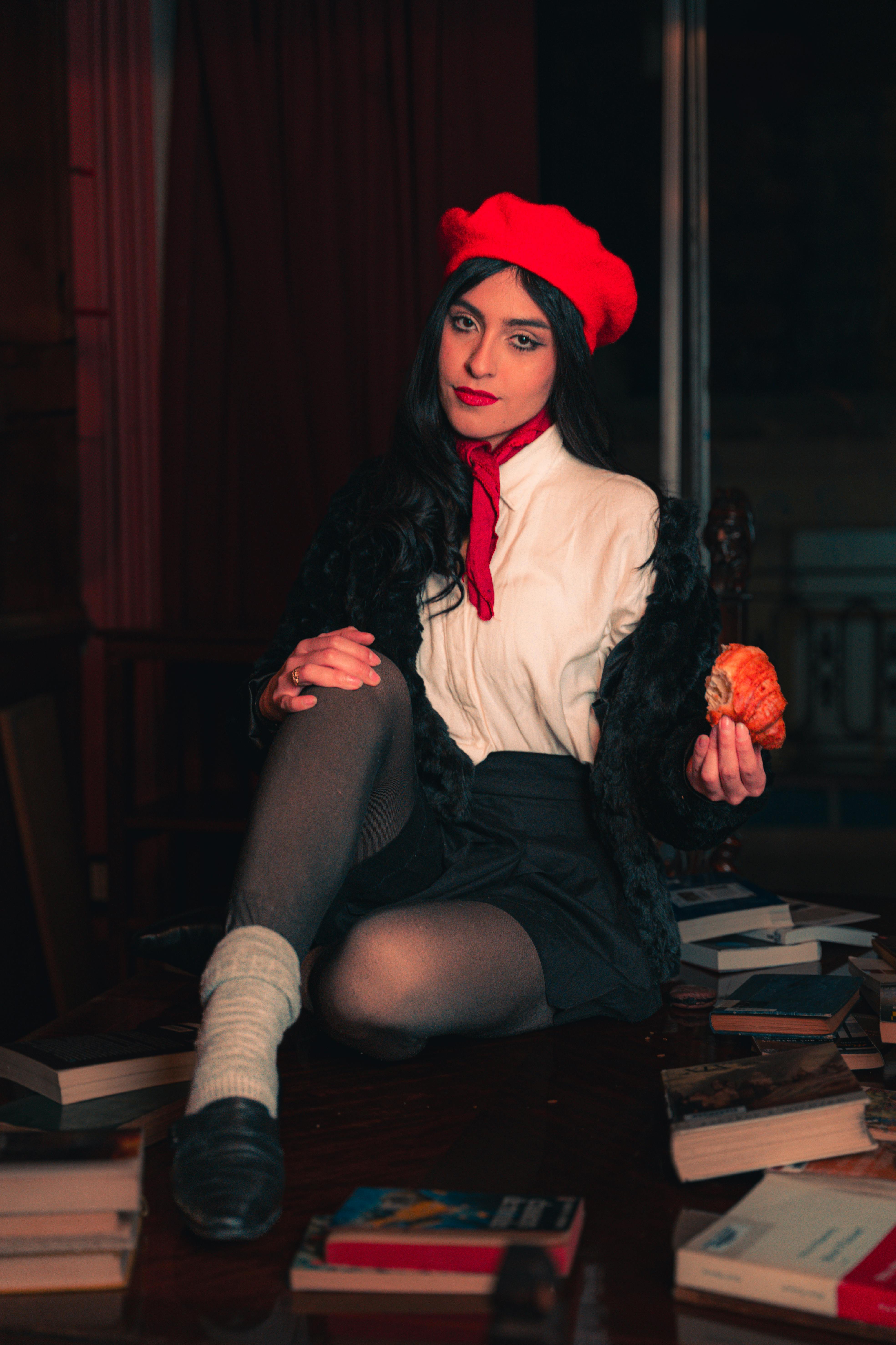 Stylish woman in a red beret sitting among books, holding an orange in a dimly lit library.
