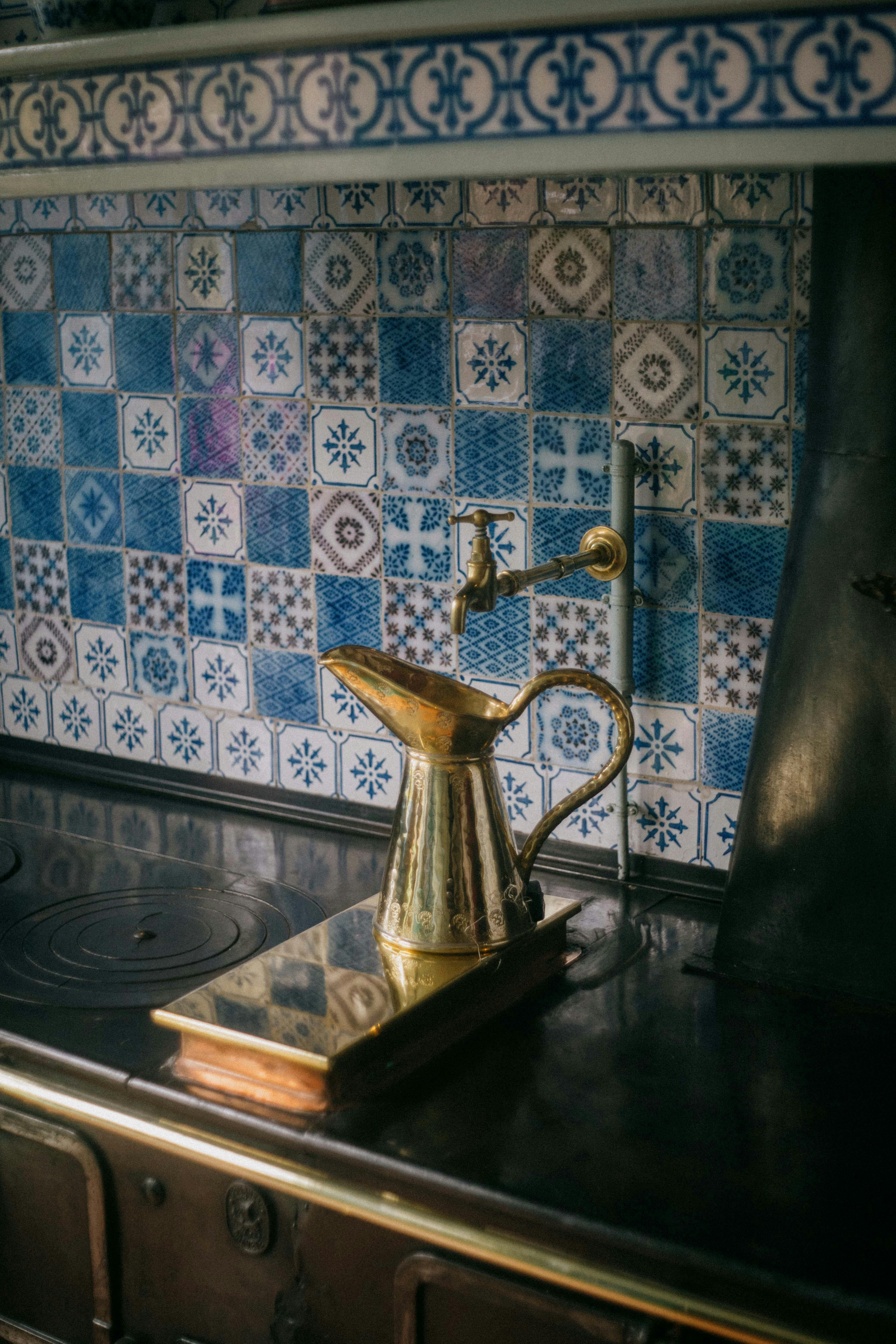 A brass kettle rests on an antique stove, set against a backdrop of intricate blue and white mosaic tiles.