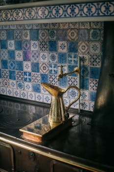 A brass kettle rests on an antique stove, set against a backdrop of intricate blue and white mosaic tiles.