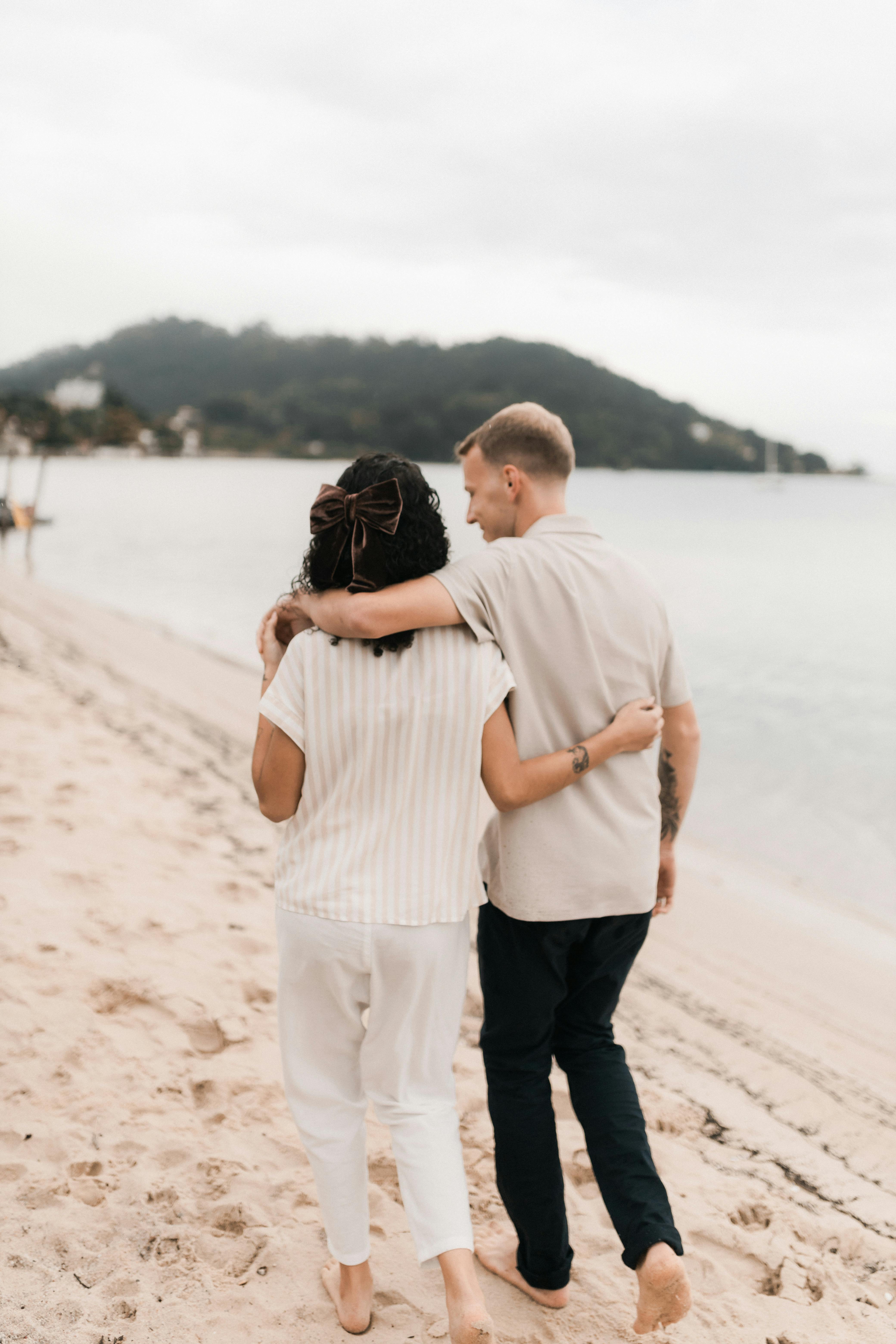 A couple walking arm in arm along a sandy beach with serene waters.