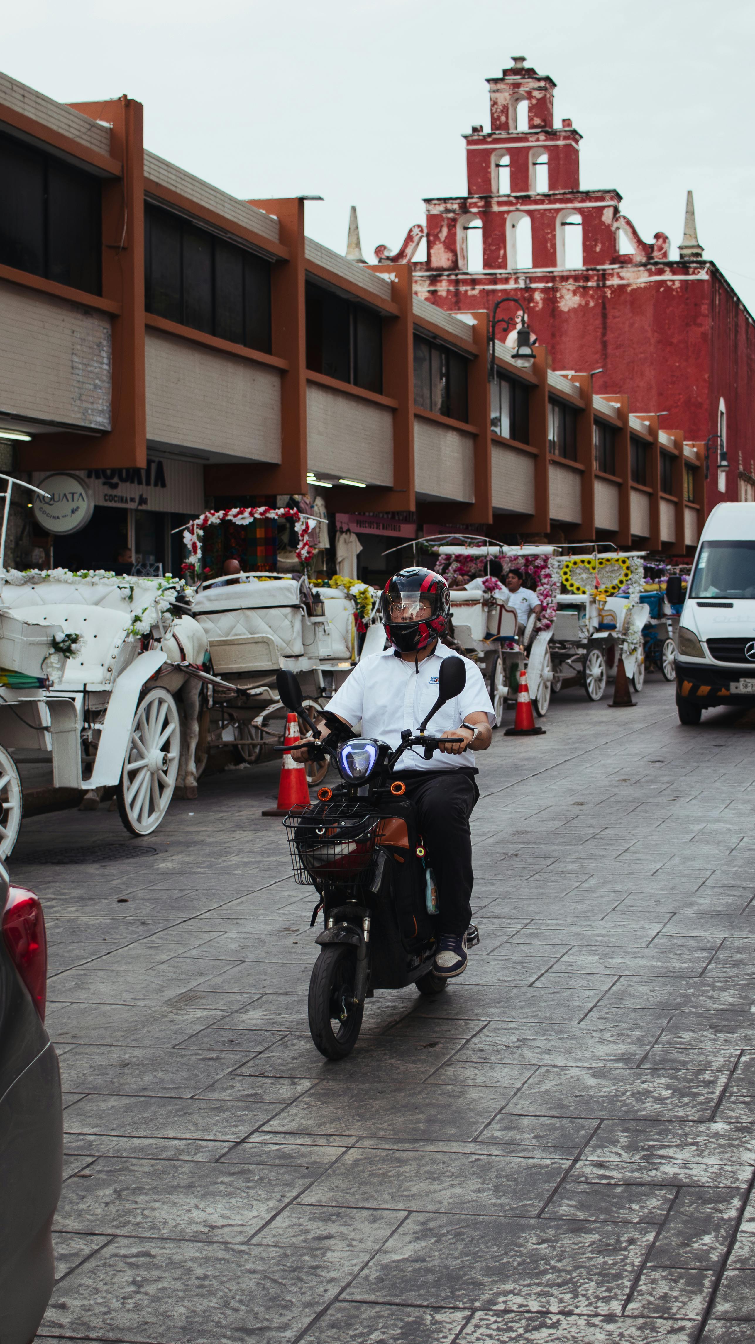 A bustling street featuring a motorbike and decorative horse carriages with a red building backdrop.