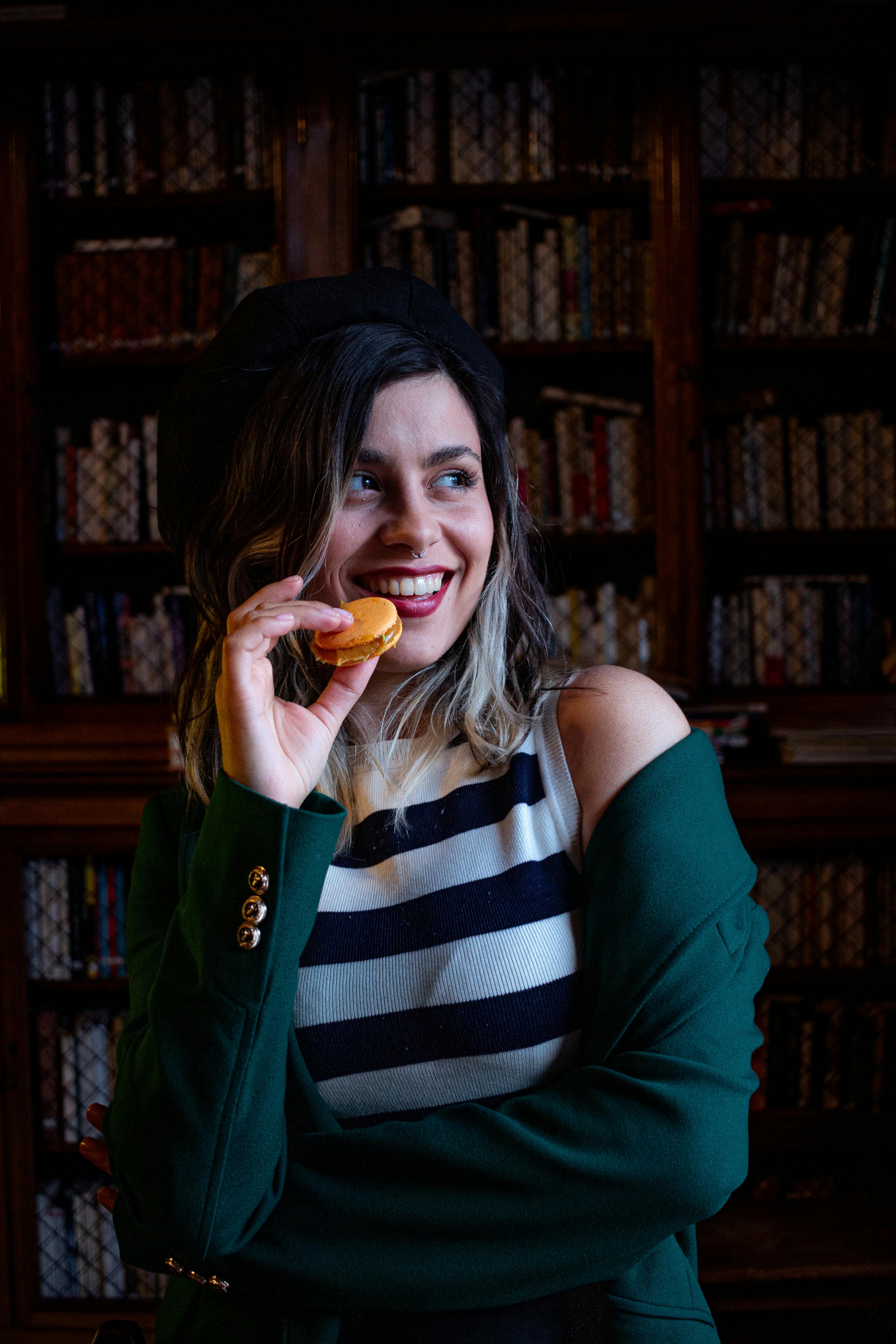 Gratis Una donna chic con un basco si gode un macaron in un'accogliente biblioteca. Un mix perfetto di moda e stile francese. Foto a disposizione