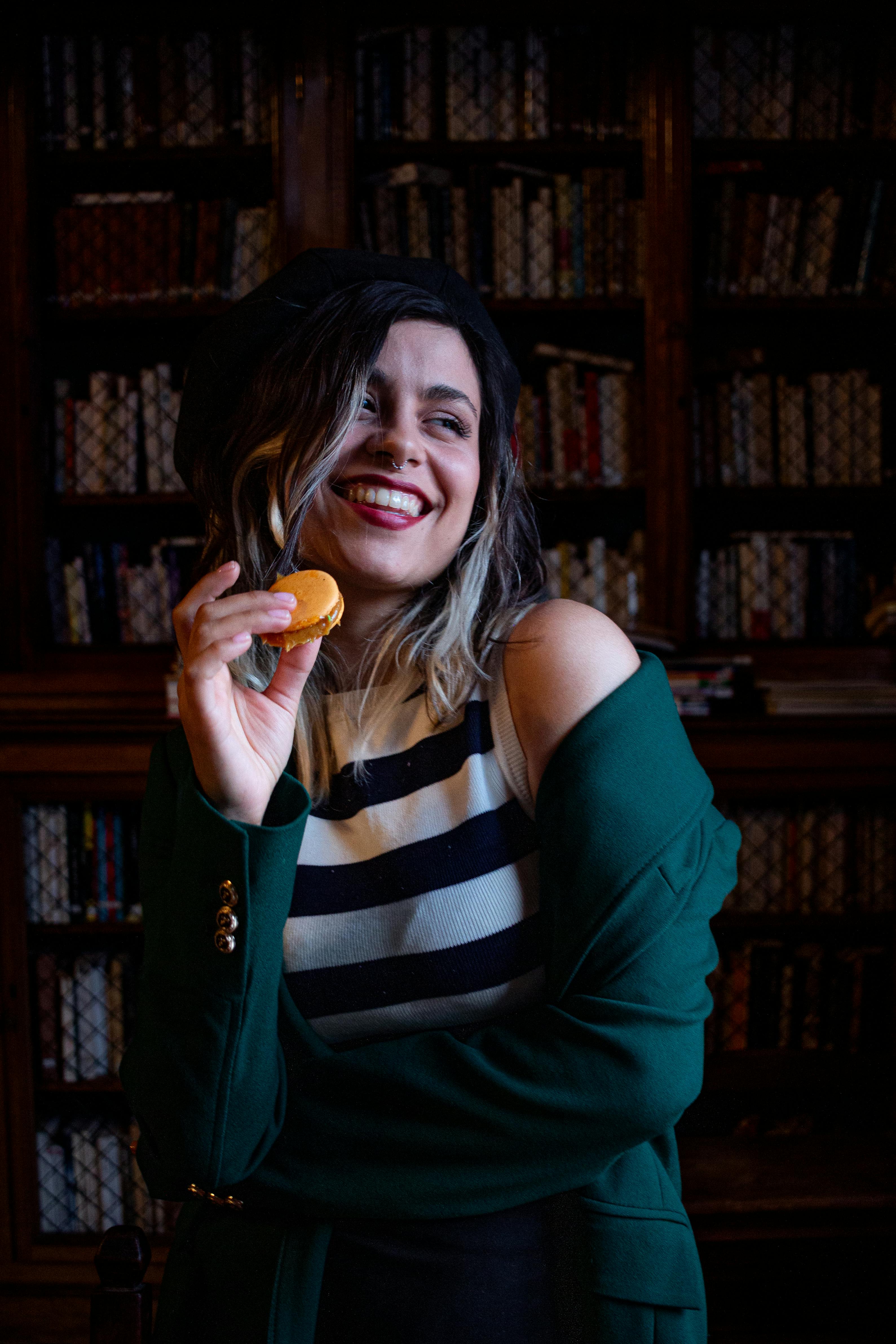 ingyenes Egy nő a könyvtárban, boldogan tartva egy macaront, sütkérezve a meleg, klasszikus hangulatban. Stockfotó