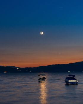 Twilight scene of boats on a tranquil lake with a crescent moon reflecting on the water.