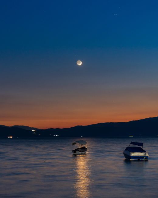 Twilight scene of boats on a tranquil lake with a crescent moon reflecting on the water.