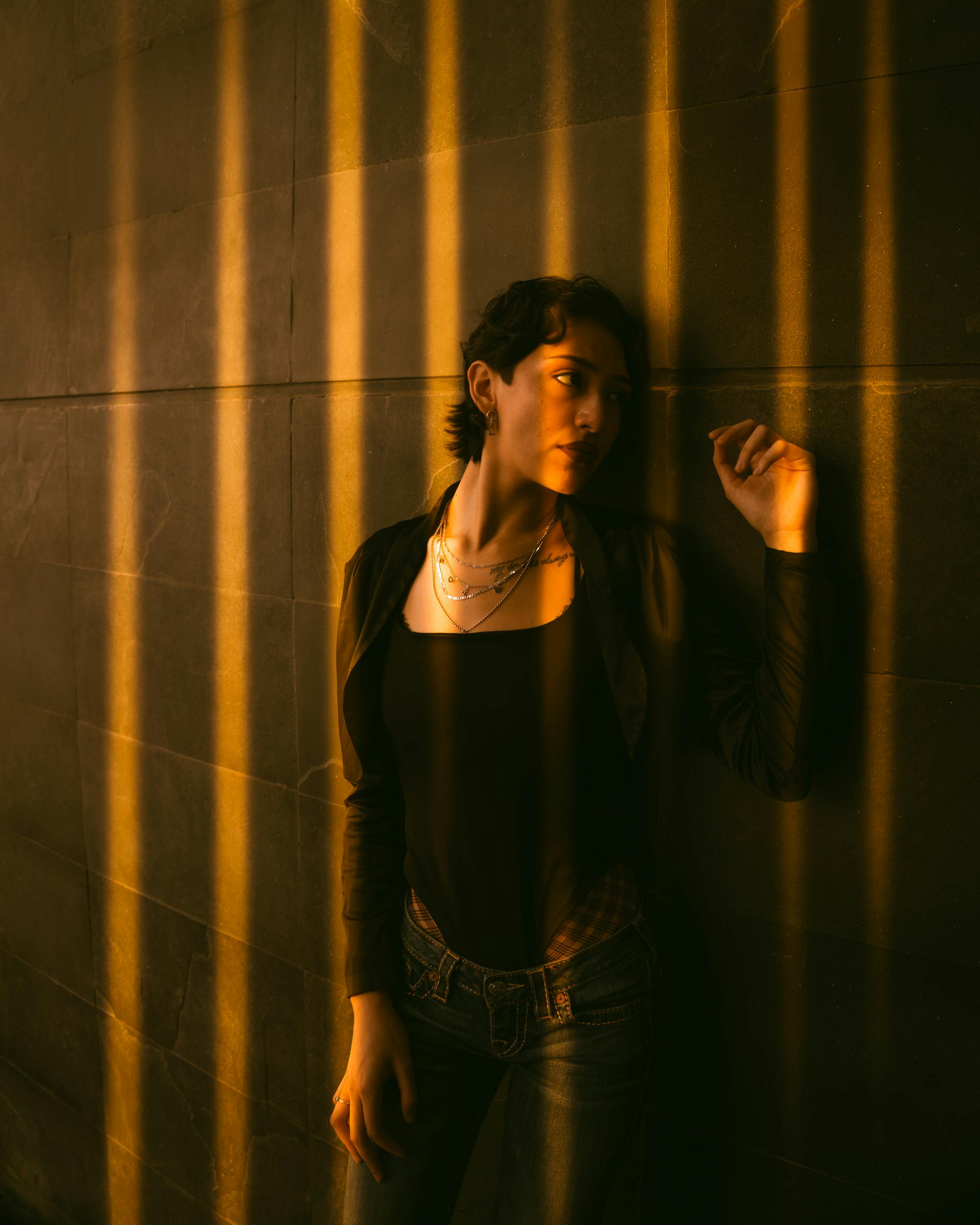 A woman in moody lighting stands against a wall with golden shadows.