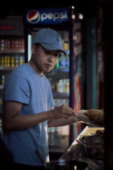 A young man in a blue shirt prepares food at a counter under a dim light, next to a beverage fridge.