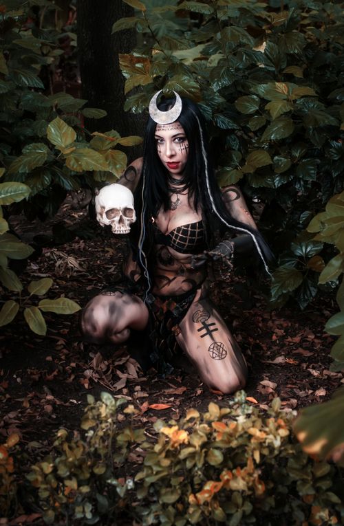Free Mystical woman dressed as a witch kneels in a dense forest, holding a skull under soft daylight. Stock Photo