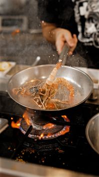 A chef expertly cooks prawns in a frying pan over a gas flame in a restaurant kitchen.