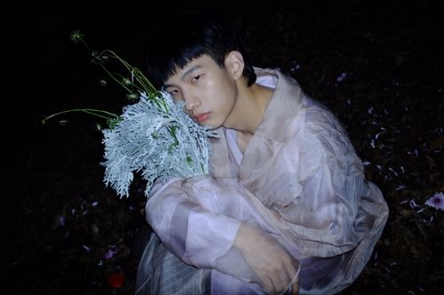 Free A contemplative young man with flowers wearing a sheer outfit in a dark setting. Stock Photo