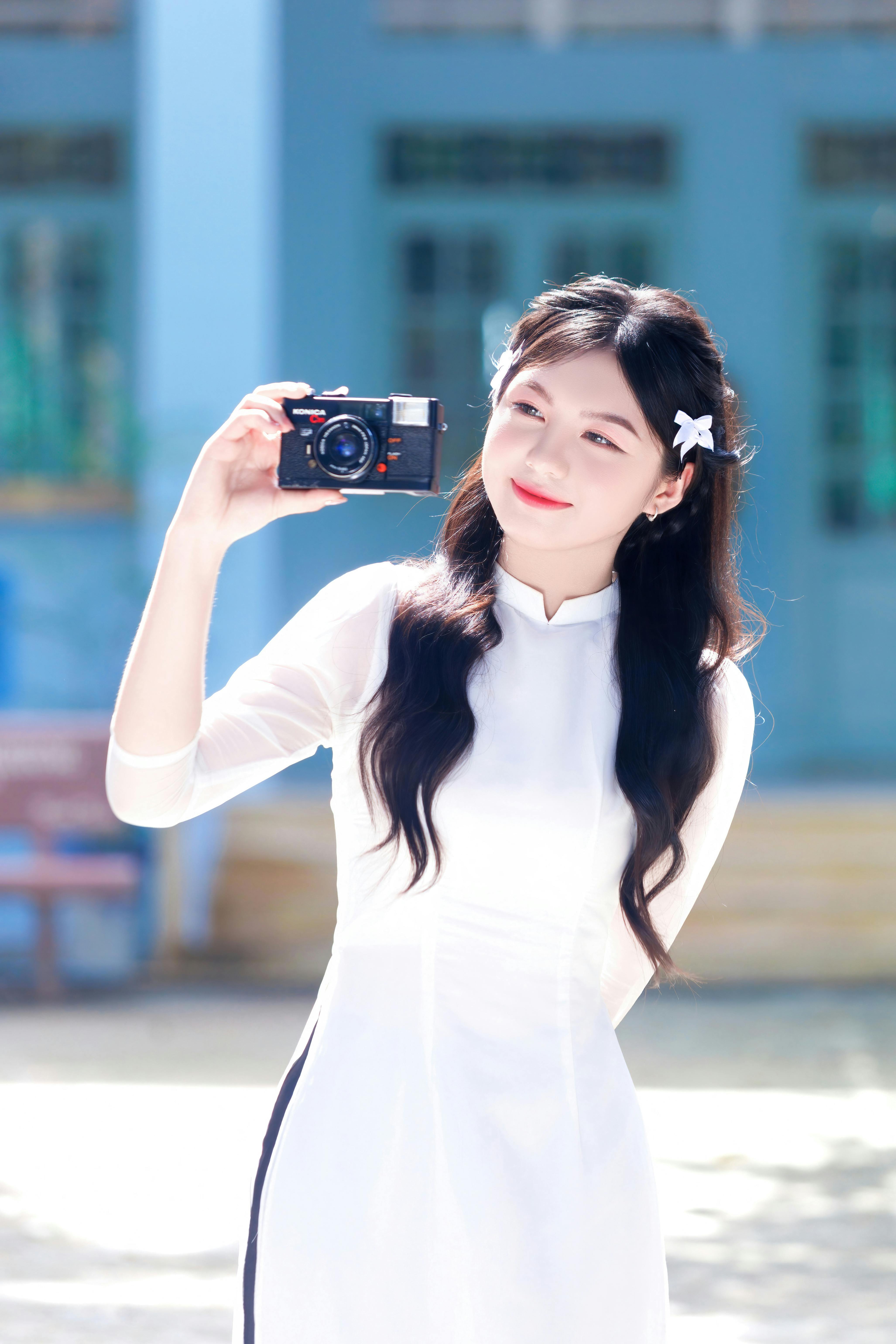 Smiling woman in a traditional white dress taking a selfie with a vintage camera outdoors.