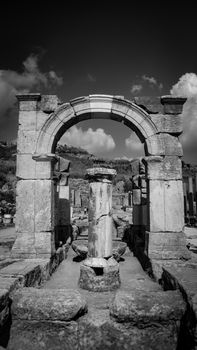 Black and white photo capturing an ancient stone archway amidst ruins under cloudy skies.