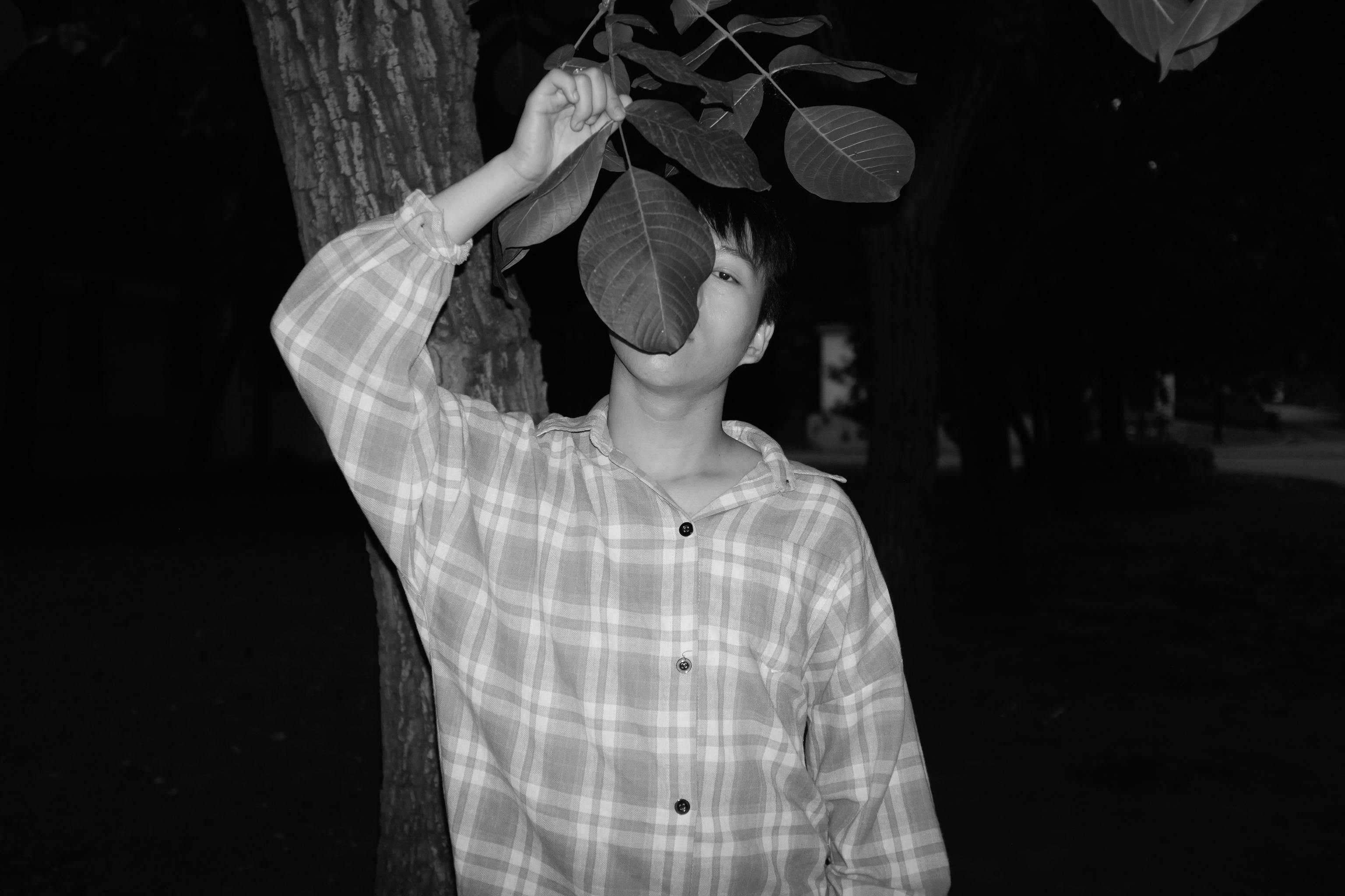 一幅黑白肖像描绘了一个人用树叶遮挡面部，站在户外的姿势