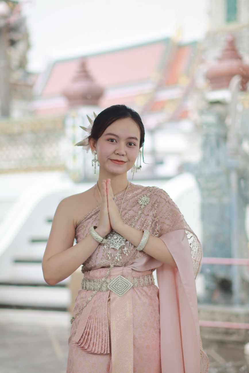 Smiling woman in ornate Thai dress posing at a temple with a traditional gesture.