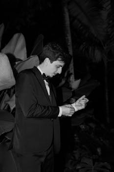 Black and white photo of a man in a tuxedo adjusting his cufflinks outdoors at night.