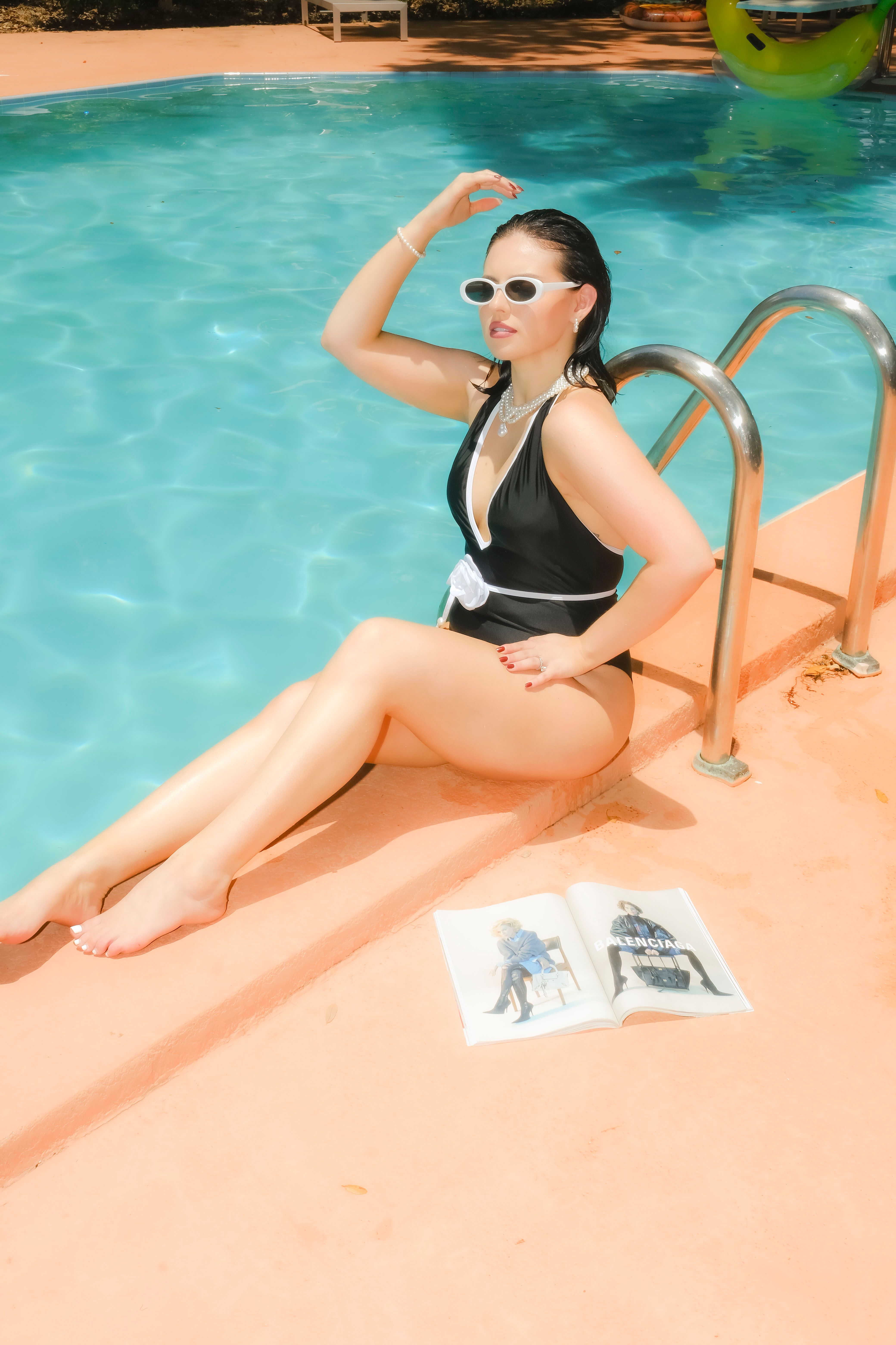 Woman in swimsuit with sunglasses by a pool enjoying a sunny day with a book.