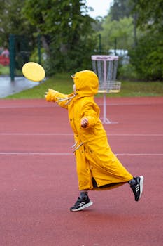 A child in a yellow raincoat plays frisbee on a rainy day in a sports field.