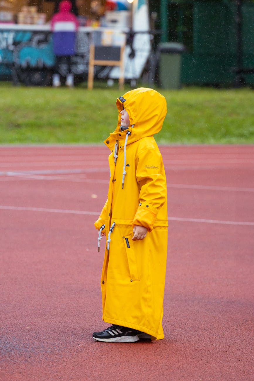 A child stands outdoors in an oversized yellow raincoat on a rainy day, adding a playful touch to the scene.