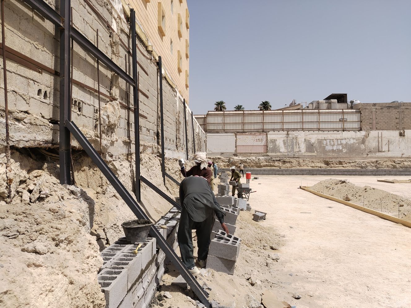 Workers building a concrete block retaining wall at a construction site under a clear sky, showcasing summer landscaping Workers building a concrete block retaining wall at a construction site under a clear sky, showcasing summer landscaping