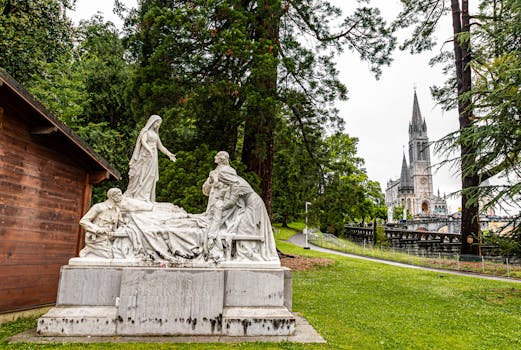 Marble statue with the Basilica of Our Lady of Lourdes in the background, amidst lush greenery.
