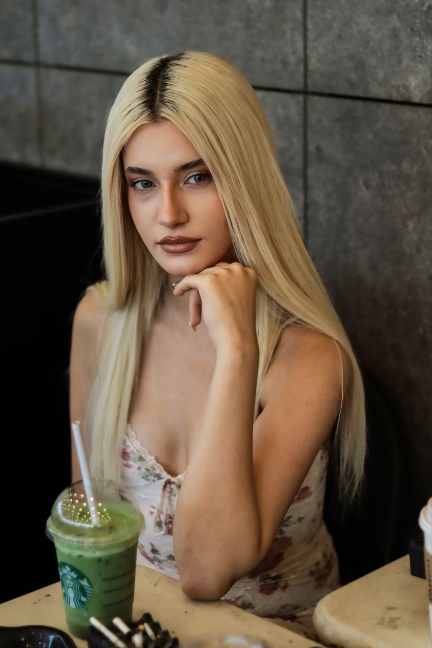 Young blonde woman seated with a green smoothie at a cafe, exuding a calm and stylish vibe.