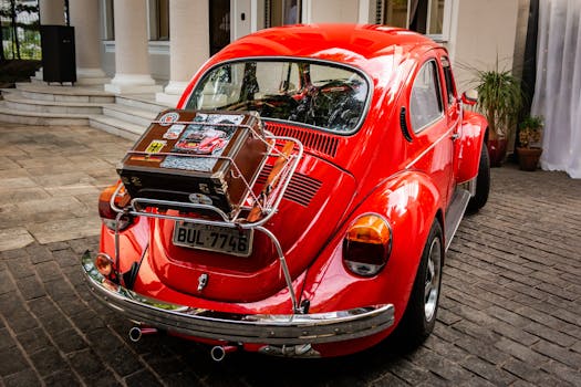 A vibrant red classic Beetle car with a vintage suitcase on its back, parked near a stylish building.